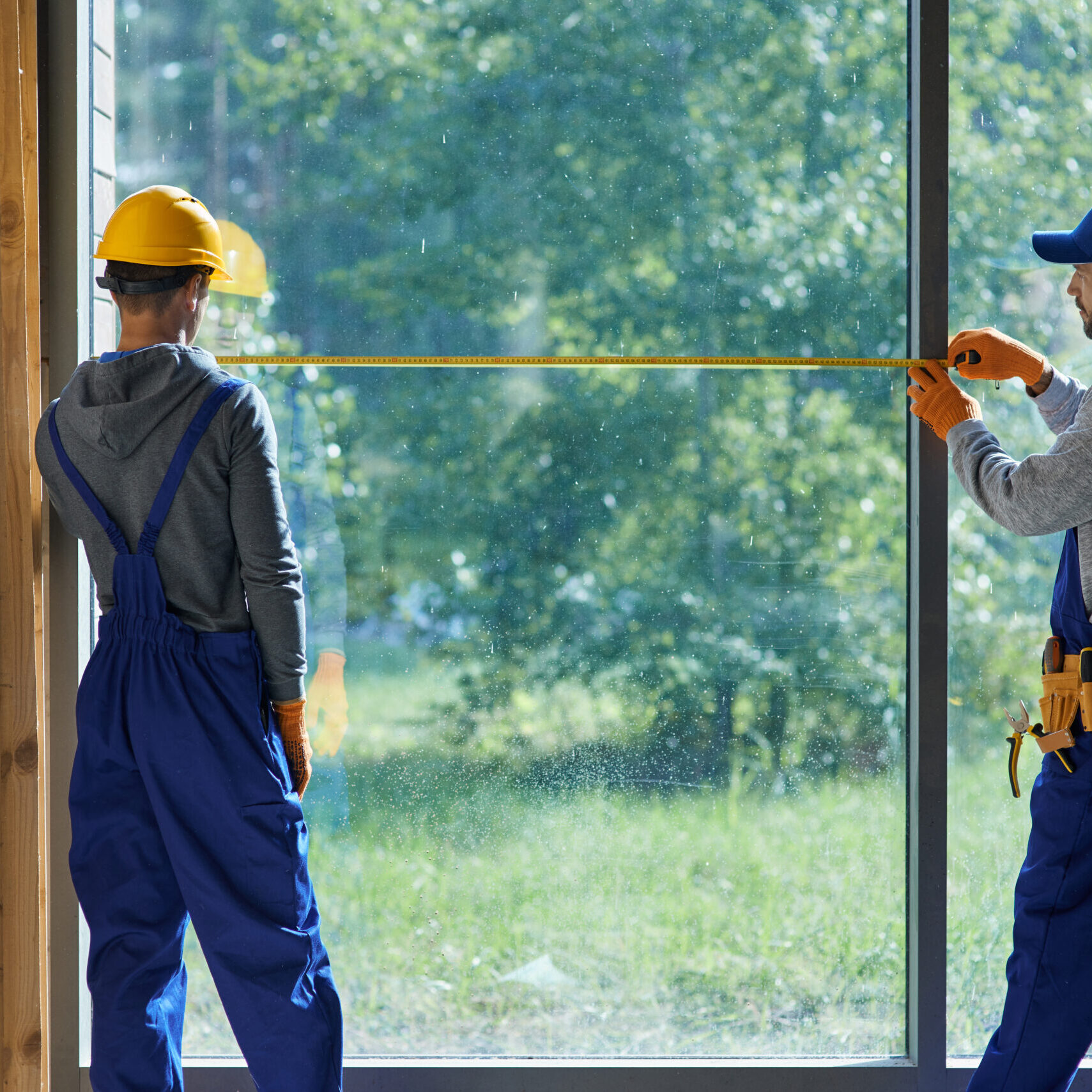 Making New Home. Rear View Of Two Young Male Builders Wearing Blue Overalls Using Measuring Tape While Working On Cottage Construction Site Making New Home. Rear View Of Two Young Male Builders Wearing Blue Overalls Using Measuring Tape While Working On Cottage Construction Site